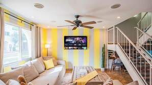 A living room with a sofa, a coffee table, a TV mounted on the wall, a ceiling fan, and a staircase. - Walk to Old Town! Pool, Arcade & Mini Golf - 6 beds (Scottsdale)