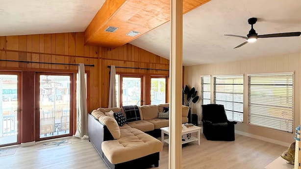 A spacious living room with a large sectional sofa, a black armchair, and a coffee table. There are large windows with blinds, a ceiling fan, and a wooden ceiling. - Spacious 3 Bedroom, 2 Bath Canal Home in the Heart of Tavernier (Tavernier)