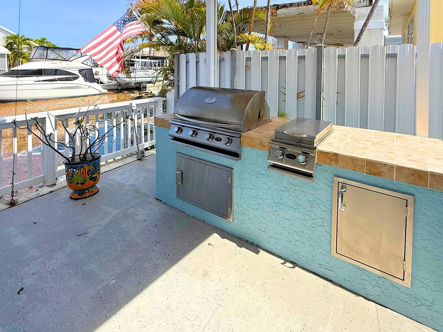 Une cuisine extérieure équipée d’un barbecue, d’un évier et d’un meuble de rangement, le tout installé devant une clôture blanche, avec une plante en pot et un drapeau américain en arrière-plan.