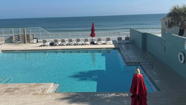 Zona de piscina con vistas al océano, sillas de descanso y un paraguas rojo.