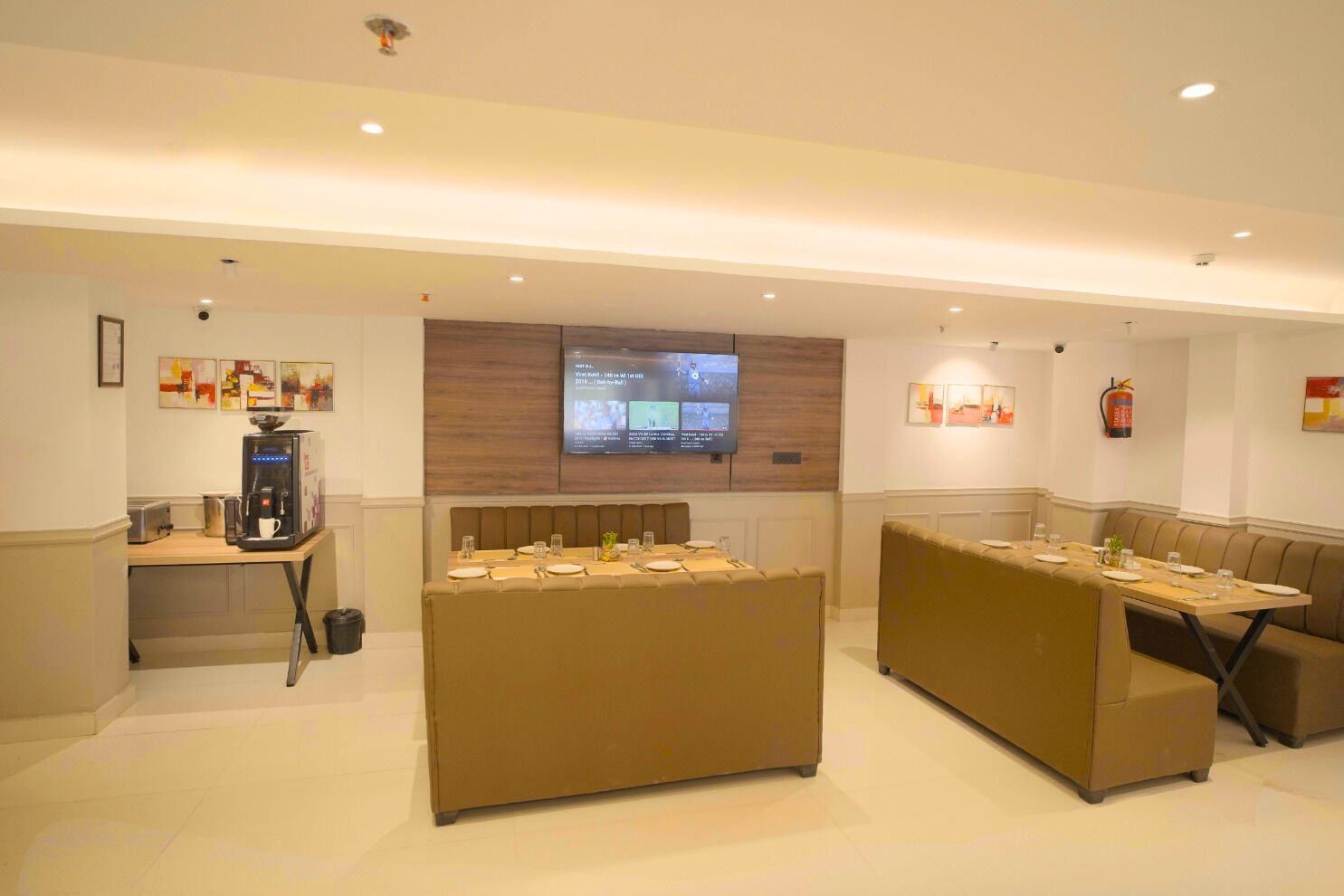A hotel lobby with a coffee station, seating areas, a TV screen, and artwork on the walls.