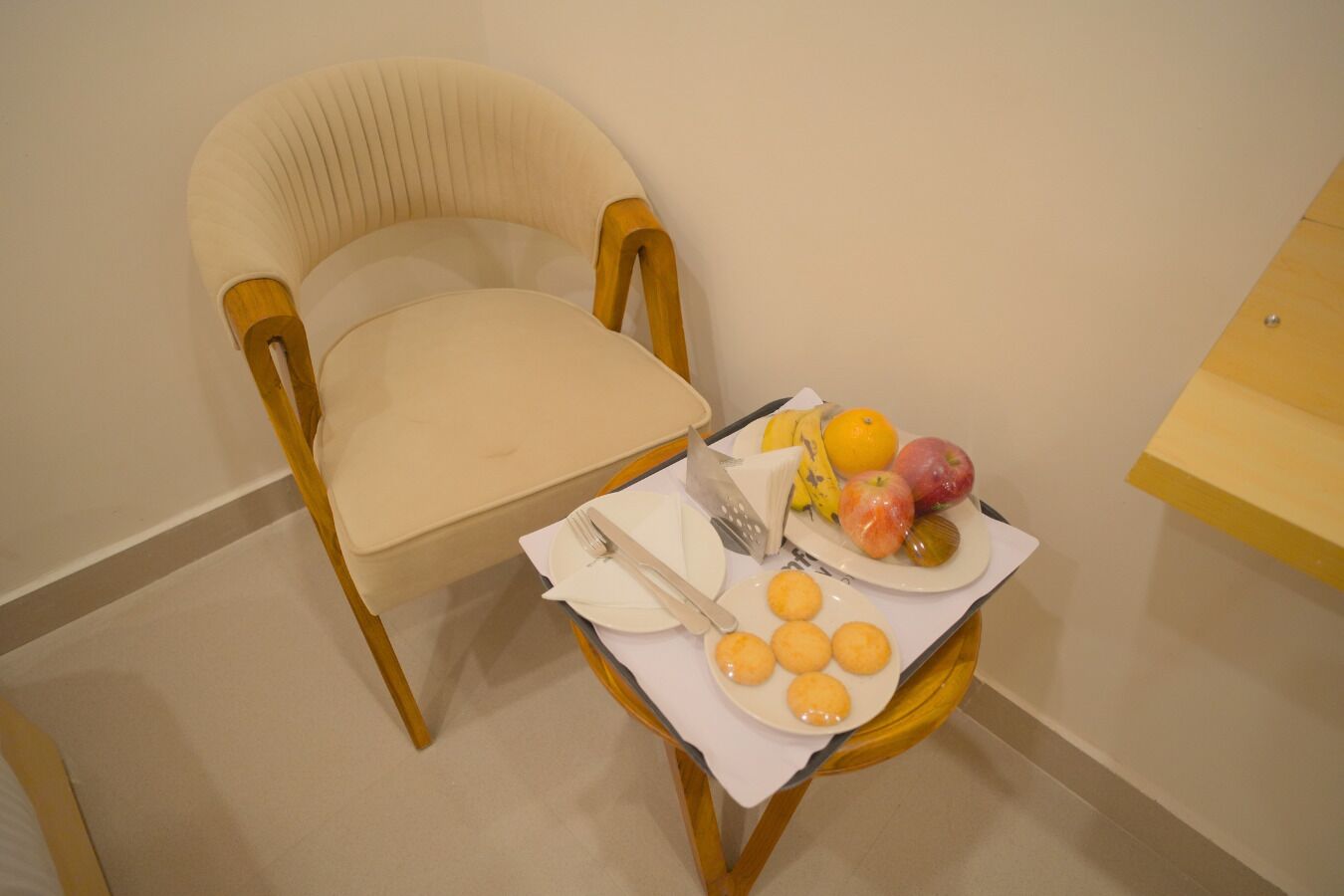 A chair with a beige cushion, a small table with a white napkin, a plate with fruits and cookies, and a knife and fork on the napkin.