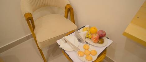A chair with a beige cushion, a small table with a white napkin, a plate with fruits and cookies, and a knife and fork on the napkin.