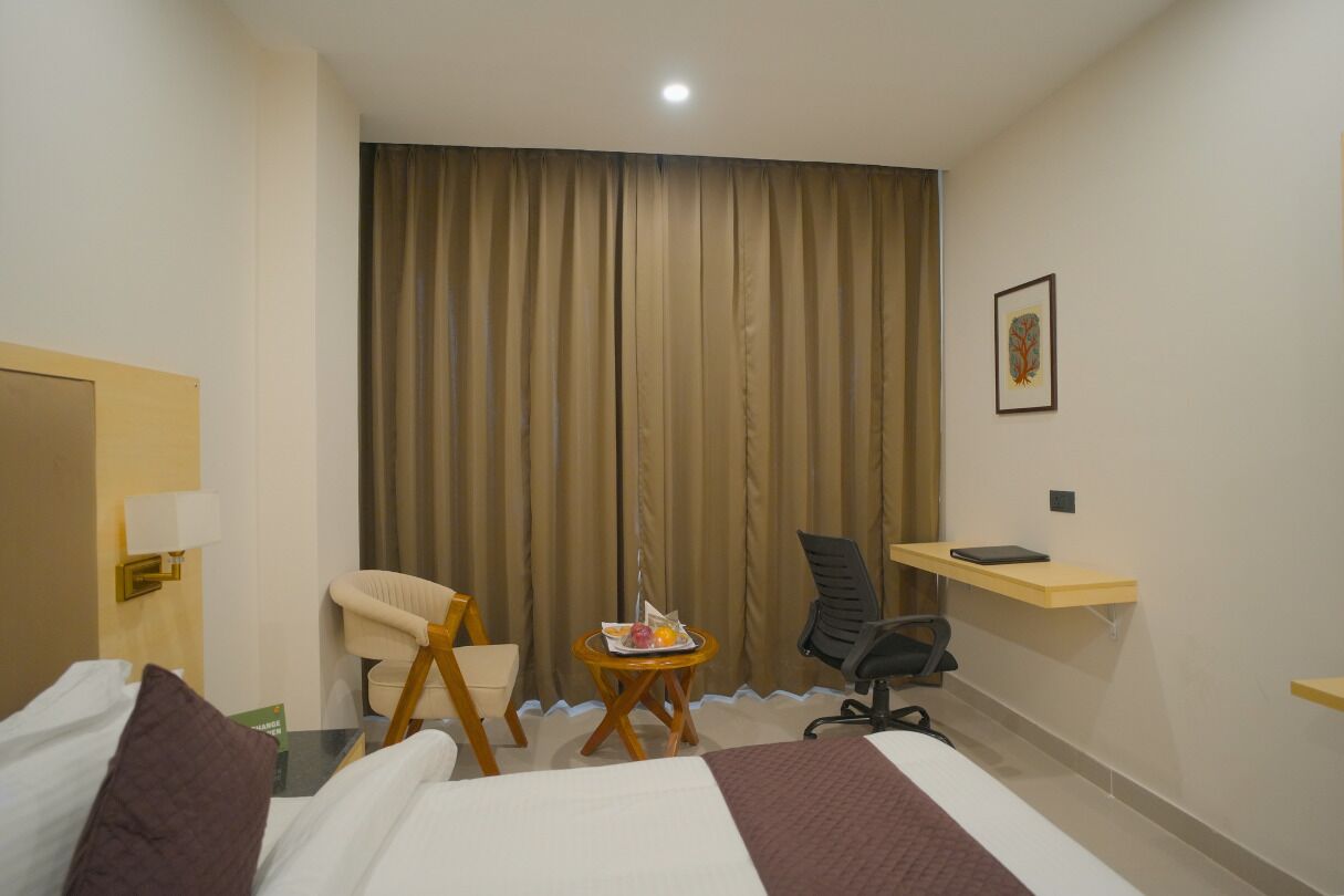 Une chambre d’hôtel avec un lit, un bureau, une chaise, une petite table sur laquelle se trouve un plateau de fruits, et une grande fenêtre avec des rideaux.
