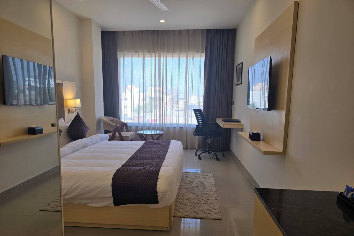 Una habitación de hotel con cama, escritorio, silla, televisor y ventana con cortinas.