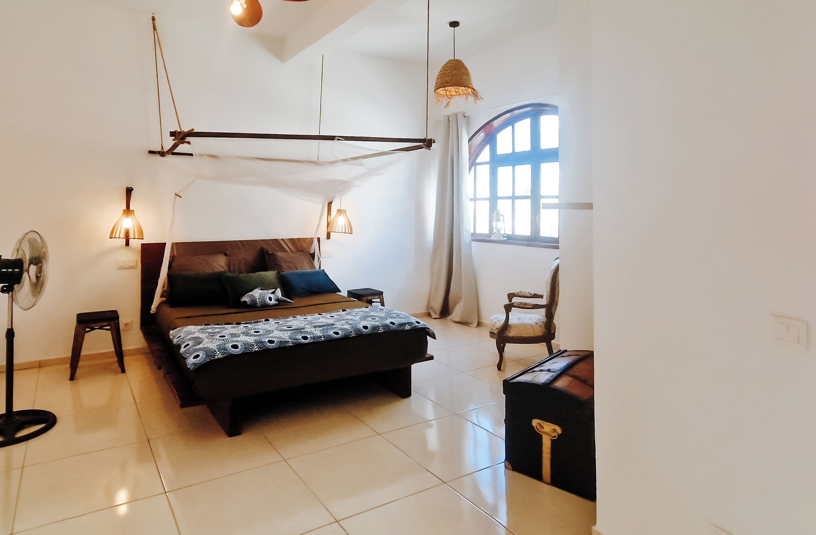 A bedroom with a bed, bedside lamps, a standing fan, a chair, and a window with curtains.
