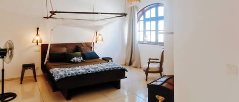 A bedroom with a bed, bedside lamps, a standing fan, a chair, and a window with curtains.