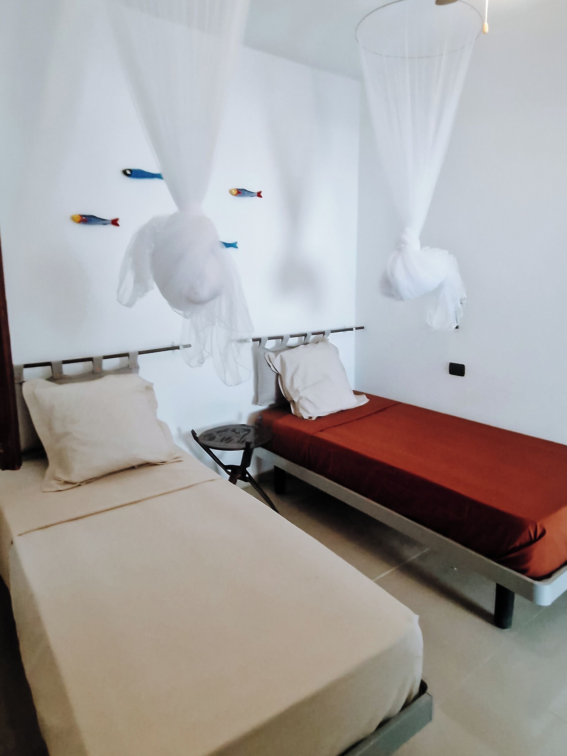 A room with two beds, one with a red mattress and the other with a white mattress. There are white curtains and a small table with a chair.