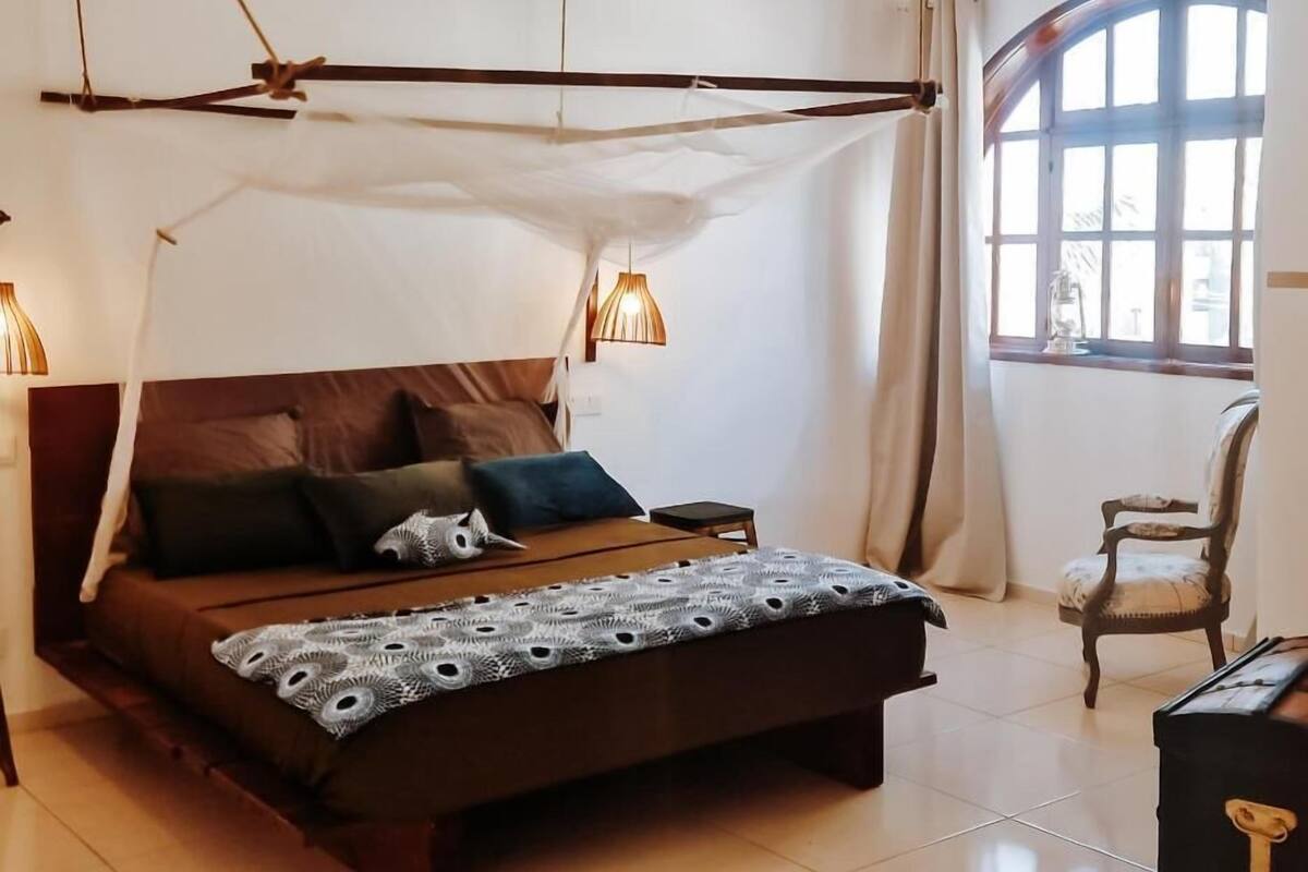 A bedroom with a four-poster bed, a wooden chair, and a window with curtains.