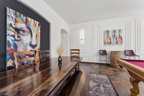 Un espace repas comprenant une table en bois, des chaises, un banc, une table de billard et deux tableaux.