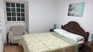 A bedroom with a bed, a chair, a nightstand with a lamp, and a window. - Caminho da Praia Apartment (Praia do Almoxarife)