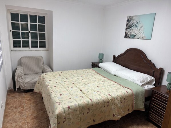 A bedroom with a bed, a chair, a nightstand with a lamp, and a window. - Caminho da Praia Apartment (Praia do Almoxarife)
