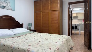 A bedroom with a wooden wardrobe, a bed with floral bedding, and a view into the kitchen area. - Caminho da Praia Apartment (Praia do Almoxarife)