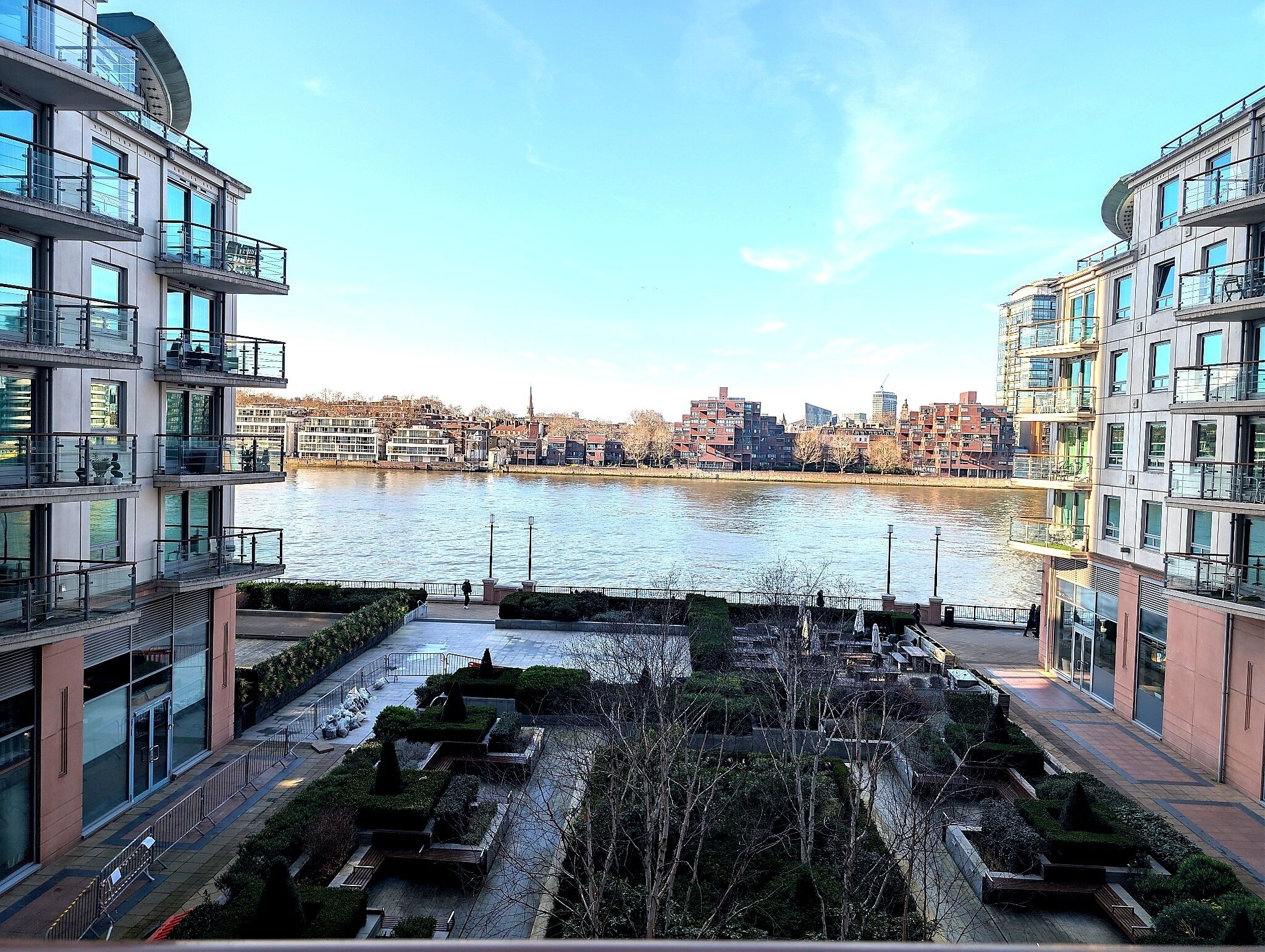 Un immeuble d’appartements moderne doté de balcons donnant sur une rivière, avec une vue sur la ville en arrière-plan et un jardin bien entretenu.