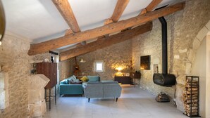 A rustic stone-walled room with exposed wooden beams, a sofa, a fireplace, and a log storage. - 18th century Provencal farmhouse - semi-detached - with exclusive pool area (Saint-Étienne-du-Grès)