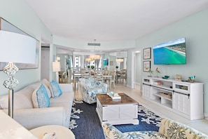 A spacious living area with a large TV, a dining area, and a kitchen in the background. - 🏖️FREE Beach Service-Pools + Hot Tub | Gulf Views | Surfside Resort #410🌊 (Miramar Beach)