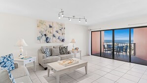 A modern living room with a grey sofa, a coffee table, and a balcony with a view of the ocean. - Gulf Coast Bliss Awaits - Reef Club 505 (Indian Rocks Beach)