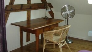 A small, cozy room with a wooden desk, a wicker chair, a standing fan, and a window providing natural light.