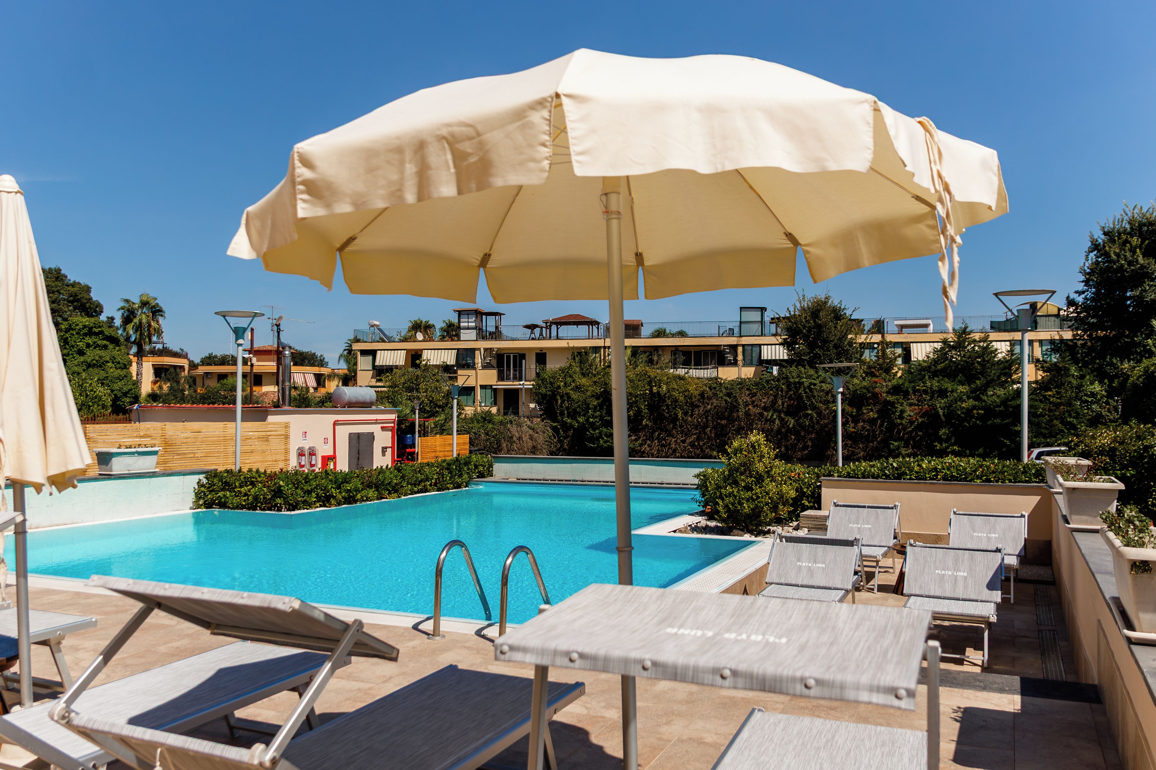 A swimming pool with lounge chairs and umbrellas, surrounded by buildings and greenery.