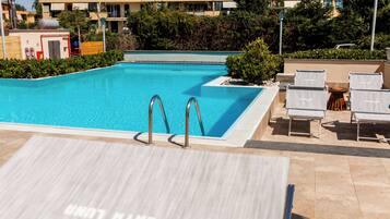 A swimming pool with a tiled deck area, lounge chairs, and a building in the background.