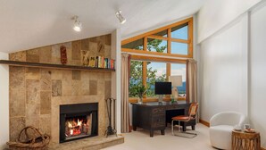 A living room with a fireplace, a desk with a computer, a chair, and a view of the outdoors through large windows.