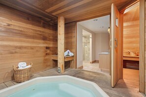 Interior de una cabaña de madera con un jacuzzi, una cesta de mimbre con toallas y una puerta de madera que da a un baño.