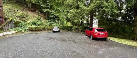 Un espace de stationnement où une voiture rouge est garée à droite et une voiture argentée est visible au loin.