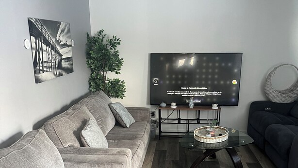 A modern living room with a grey sectional sofa, a grey armchair, a coffee table, and a flat-screen TV mounted on the wall. - Soothing 2-bedroom house with AC in charming 3 mins from Dearborn Hts (Inkster)