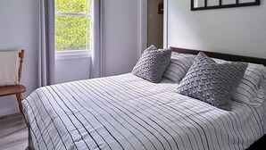 A neatly made bed with striped bedding and two pillows, a wooden chair with a white cushion, and a window with curtains. - Whimsical Waterside Cottage (Sharbot Lake)