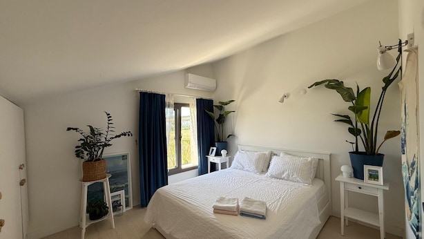Un dormitorio con una cama grande, una ventana con cortinas, una planta en maceta y una mesita de noche con una lámpara.