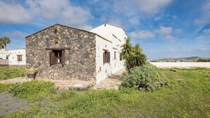 A stone building with a white upper section, a small porch, and a view of the beach and hills in the background. - Home2Book Casa María Rustic Escape in Villaverde (Villaverde)
