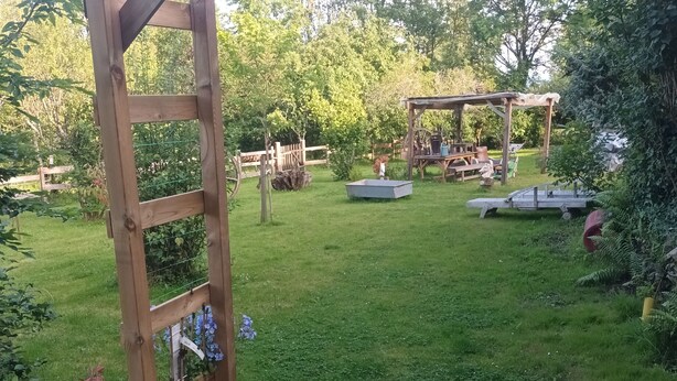 A wooden pergola structure in a garden with a grassy lawn, surrounded by trees and plants.