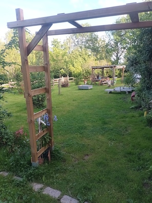 A wooden pergola structure in a garden with a grassy lawn, surrounded by trees and plants.
