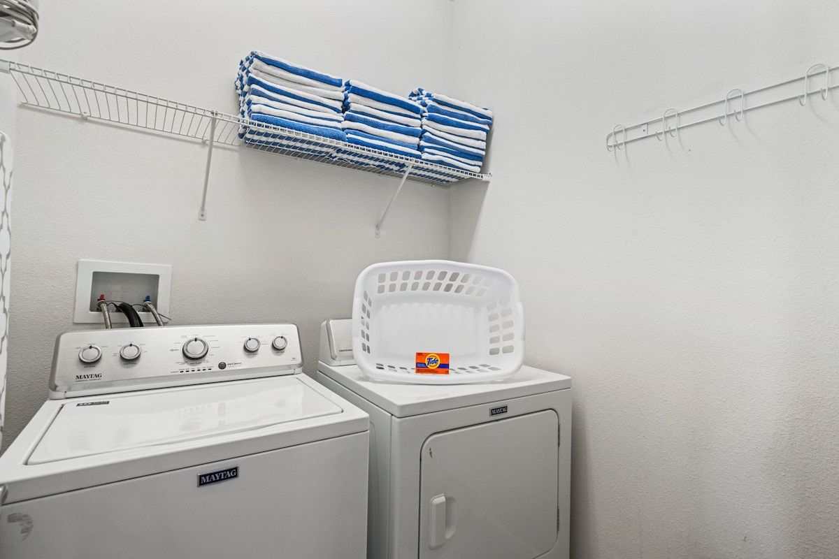 A laundry area with a Maytag washer and dryer, a white basket, and a shelf with folded towels.
