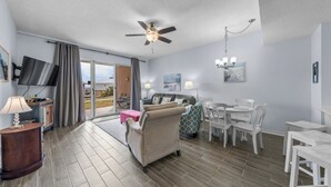 Ein geräumiges Wohnzimmer mit Deckenventilator, einem Flachbildfernseher, einer Essecke mit weißen Stühlen und einem großen Fenster mit Blick ins Freie.