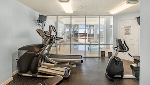 Ein Fitnessraum mit Laufbändern, Crosstrainern und einem stationären Fahrrad. Es gibt ein großes Fenster mit Blick auf einen Poolbereich.