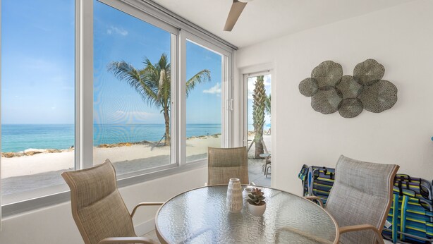Quarto com mesa de vidro, cadeiras de vime, um detalhe decorativo na parede e vista para a praia e oceano através de amplas janelas.