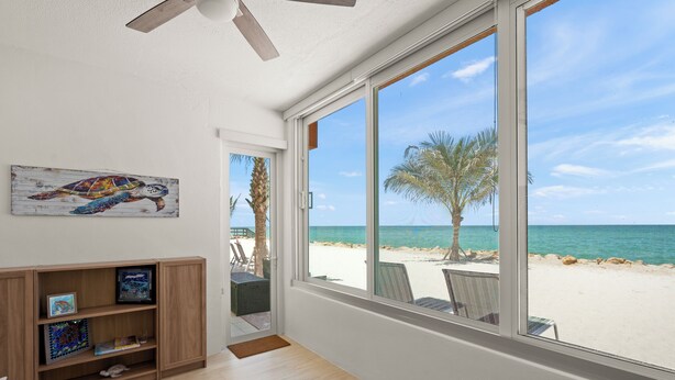 A room with a large window overlooking a beach, a ceiling fan, a wooden bookshelf, and a door leading outside. - Your Gulf-Front Getaway Awaits (Siesta Key)