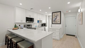 A modern kitchen with white cabinetry, a central island with stools, and a large painting on the wall. - Your Gulf-Front Getaway Awaits (Siesta Key)