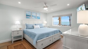 A bedroom with a bed, nightstands, a ceiling fan, a window with a view, and artwork on the wall. - Gulf-Front Serenity at Fisherman’s Cove (Siesta Key)