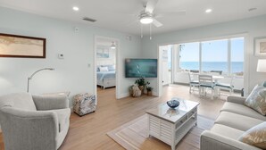 A spacious living room with a flat-screen TV, a ceiling fan, and a view of the ocean through large windows. - Gulf-Front Serenity at Fisherman’s Cove (Siesta Key)
