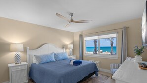 Een slaapkamer met een bed, nachtkastjes, een plafondventilator, een raam met uitzicht op het strand en een bureau met een stoel.