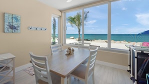 Een kamer met een houten tafel, witte stoelen, een plafondventilator, een groot raam met uitzicht op het strand en een klein plankje met spullen.