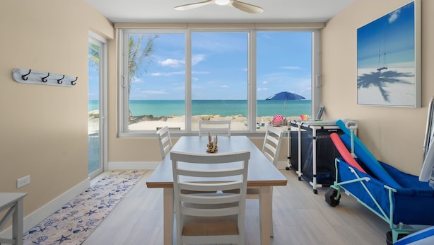 Een kamer met een eettafel, stoelen, een plafondventilator, een groot raam met uitzicht op het strand en een trolley met strandhanddoeken.