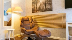 A modern room with a leather armchair and ottoman, a standing lamp, a large forest-themed painting, and a wooden slatted wall panel. - Rein Klassik Hotel (Bad Pyrmont)