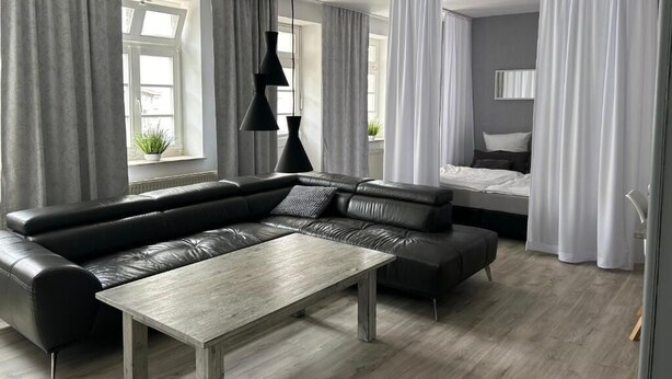 A modern hotel room with a sectional sofa, a wooden coffee table, a bed with white curtains, and a view of the outdoors. - Rein Klassik Hotel (Bad Pyrmont)