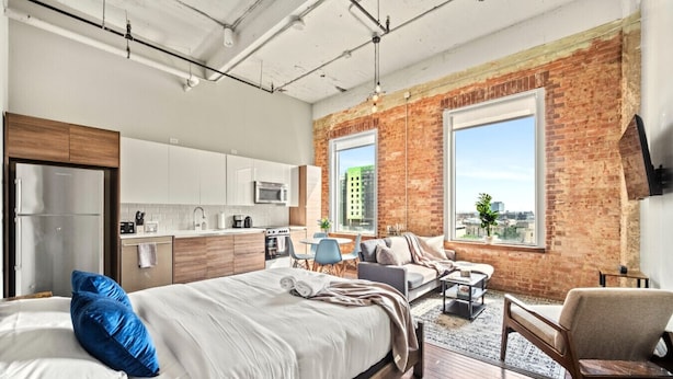 Een moderne slaapkamer met een groot bed, een zithoek met een bank en fauteuils, een keuken met witte kasten en een bakstenen muur met grote ramen.