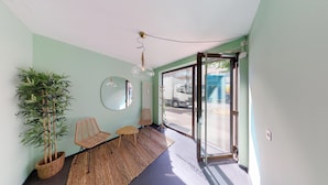 A hallway with a large potted bamboo plant, a wicker chair, a round mirror, and a ceiling light fixture. - BASELLODGING Schönau Serviced Living (Basel)