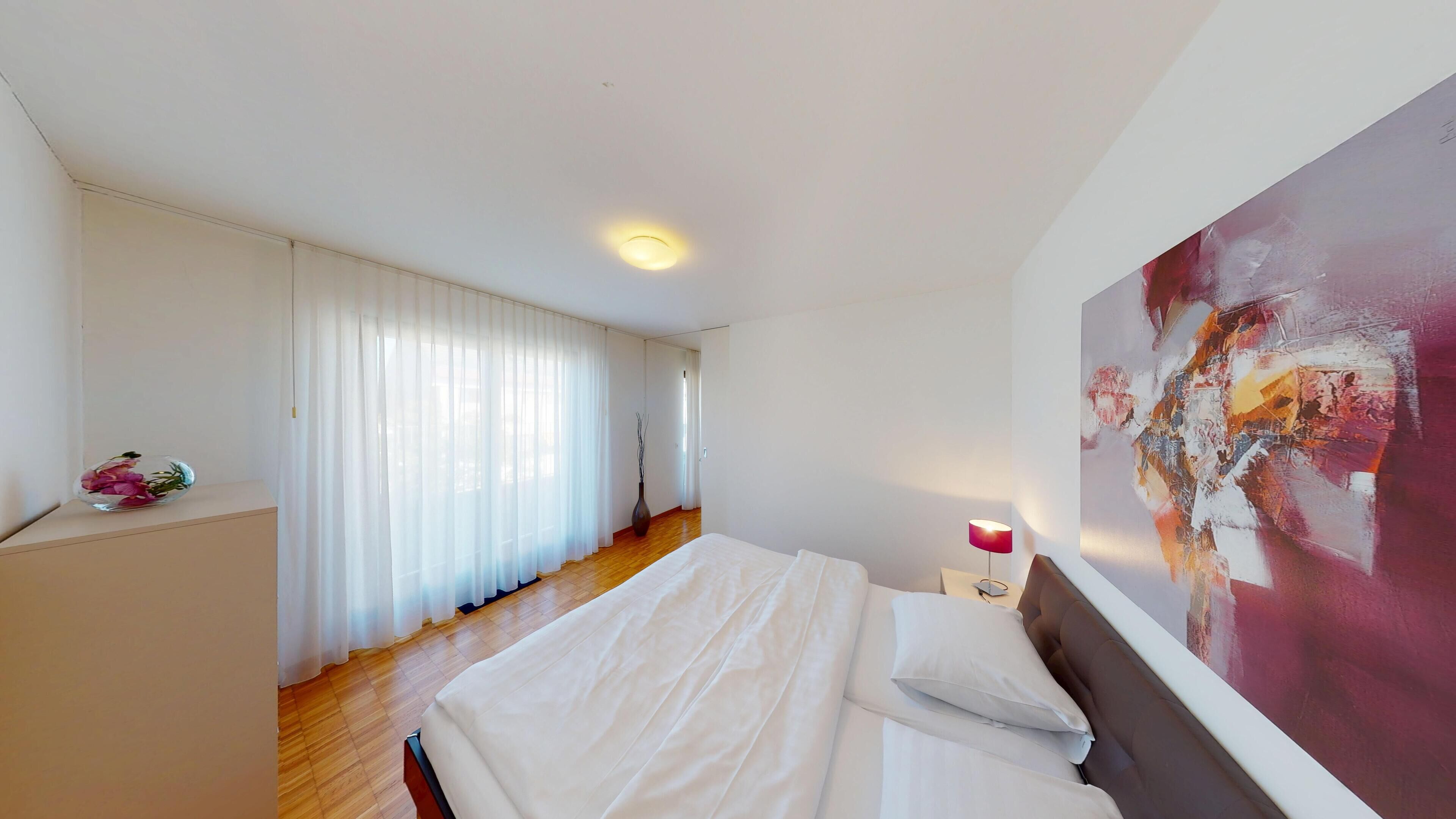 Un dormitorio con cama, una mesita de noche con lámpara, un cuadro en la pared y una ventana con cortinas traslúcidas.