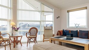 A spacious room with a large window, a wooden table, a cushioned bench, wicker chairs, and a small table with a lamp and flowers. - 5 Star Holiday Home in Lokken (Lokken)